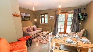 A cozy living room with a sofa, a bright orange chair, a wooden dining table set for four, and a large window with curtains. - Bwthyn Canol (Haverfordwest)