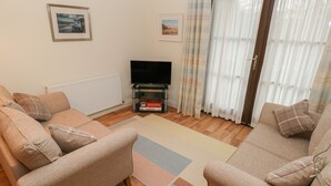 A living room with two beige sofas, a flat-screen TV on a stand, a radiator, and a window with curtains. - 2 Chandlers Yard (Burry Port)