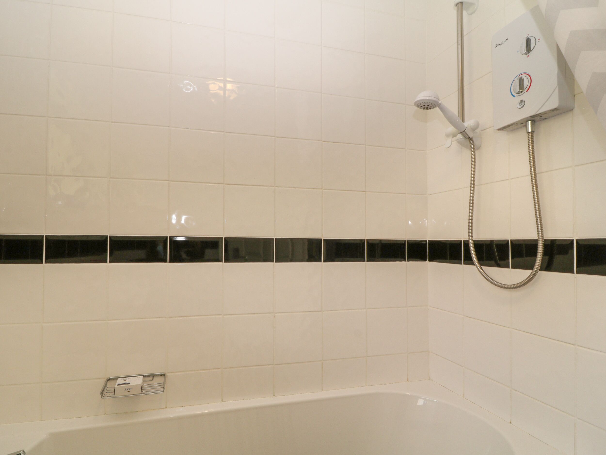 A bathroom with white tiles, a black-tile border, a bathtub, and a shower control panel.