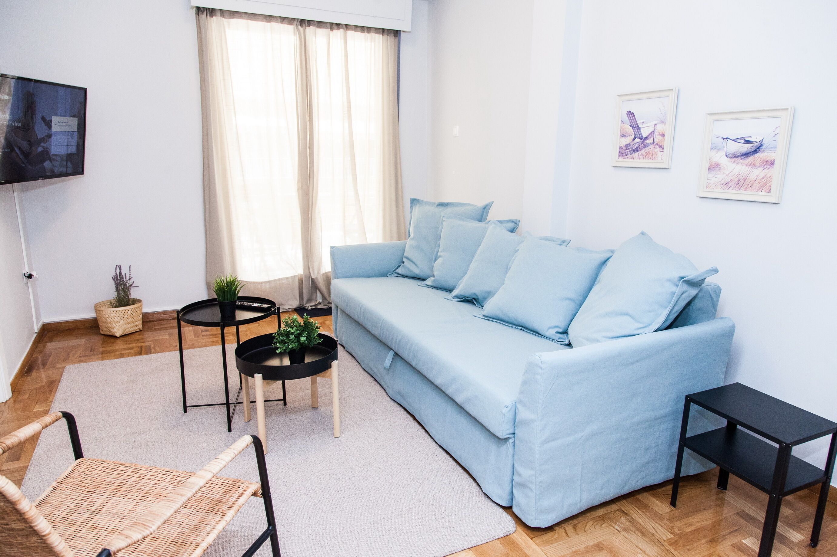 A living room with a light blue sofa, a television, two small tables, a wicker chair, and a window with curtains.