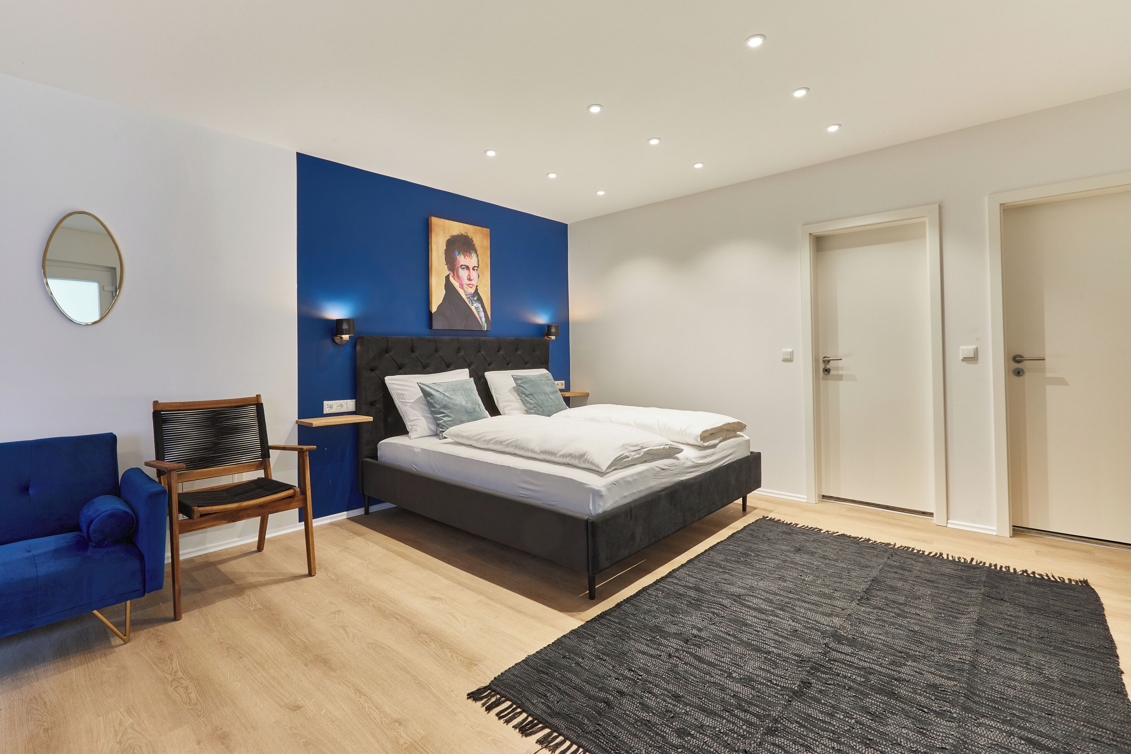 A modern bedroom with a bed, a blue wall, a painting, a mirror, and a door.