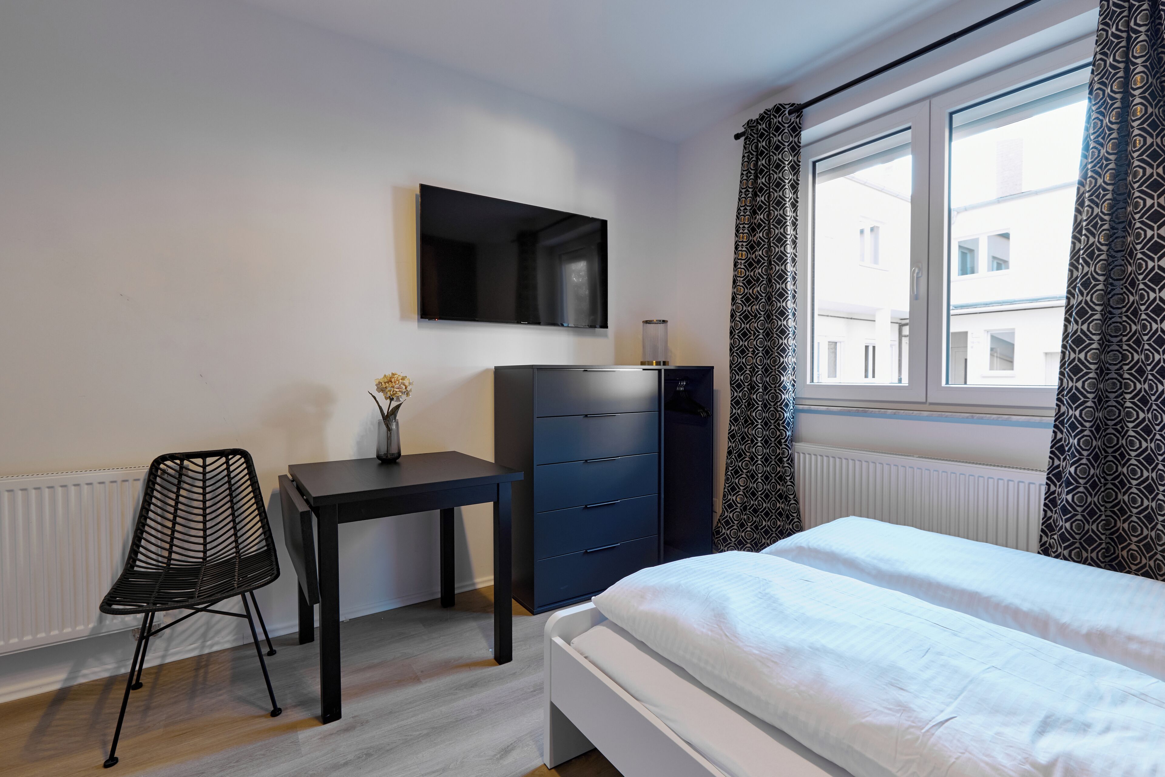 Une chambre à coucher comprenant un lit, un bureau avec une chaise, une commode, un téléviseur et une fenêtre avec des rideaux à motifs.