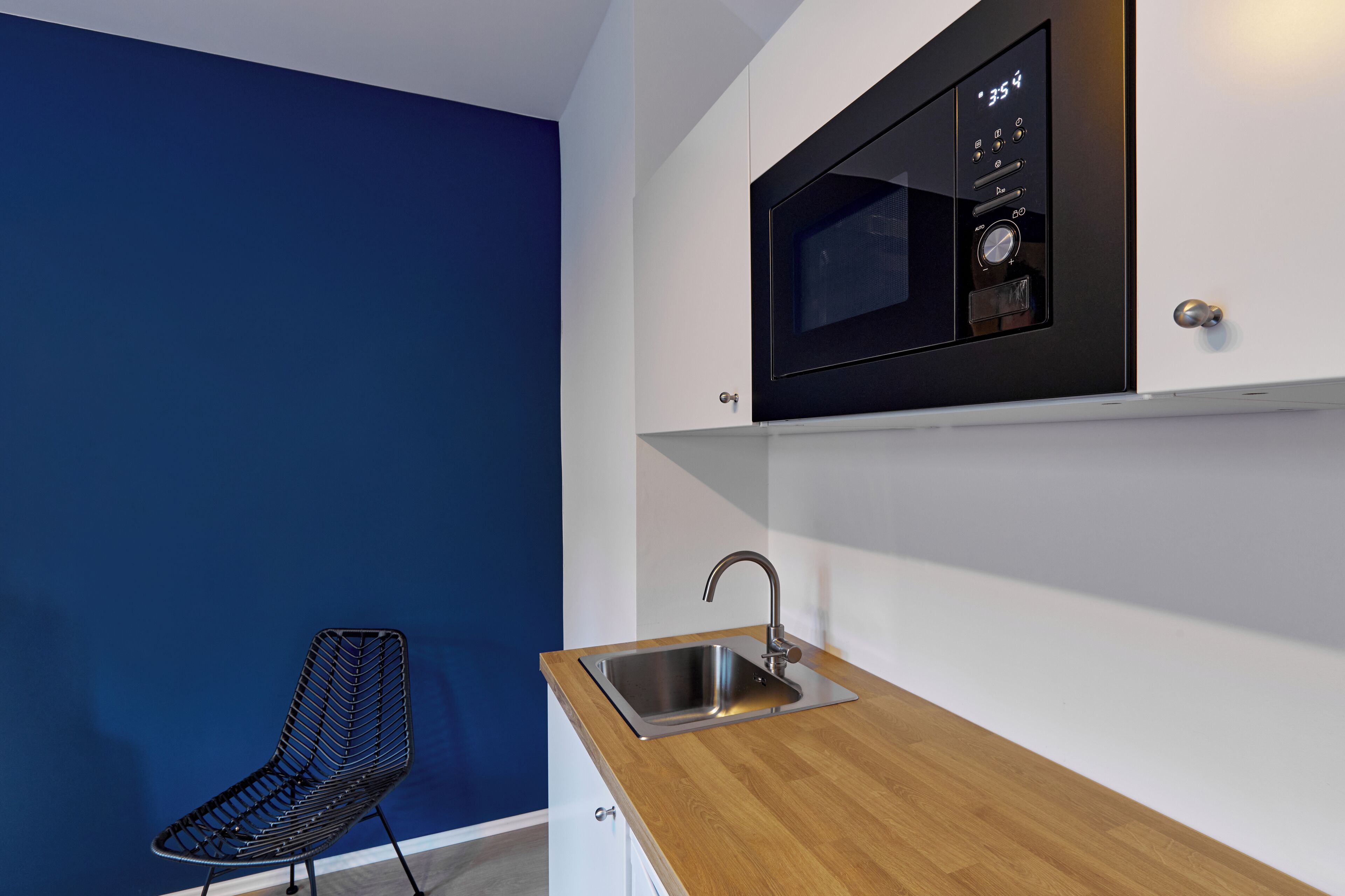 Una cocina compacta con microondas integrado, un lavamanos de acero inoxidable y una silla moderna.