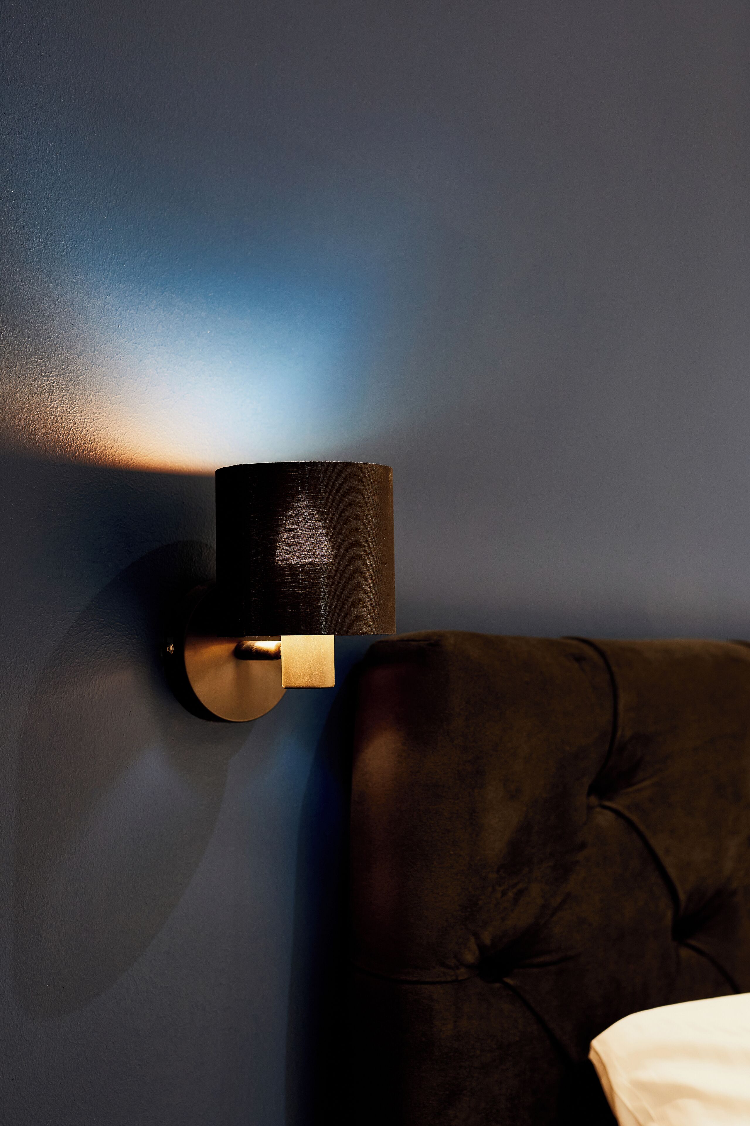 A wall-mounted light fixture with a cylindrical shade, positioned next to a dark-colored upholstered headboard.