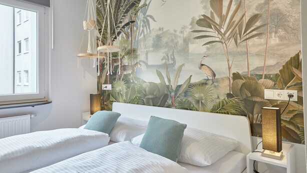 A hotel room with two beds, a mural of a tropical scene, and a window with a view of buildings. - Humboldt41 (Nuremberg)