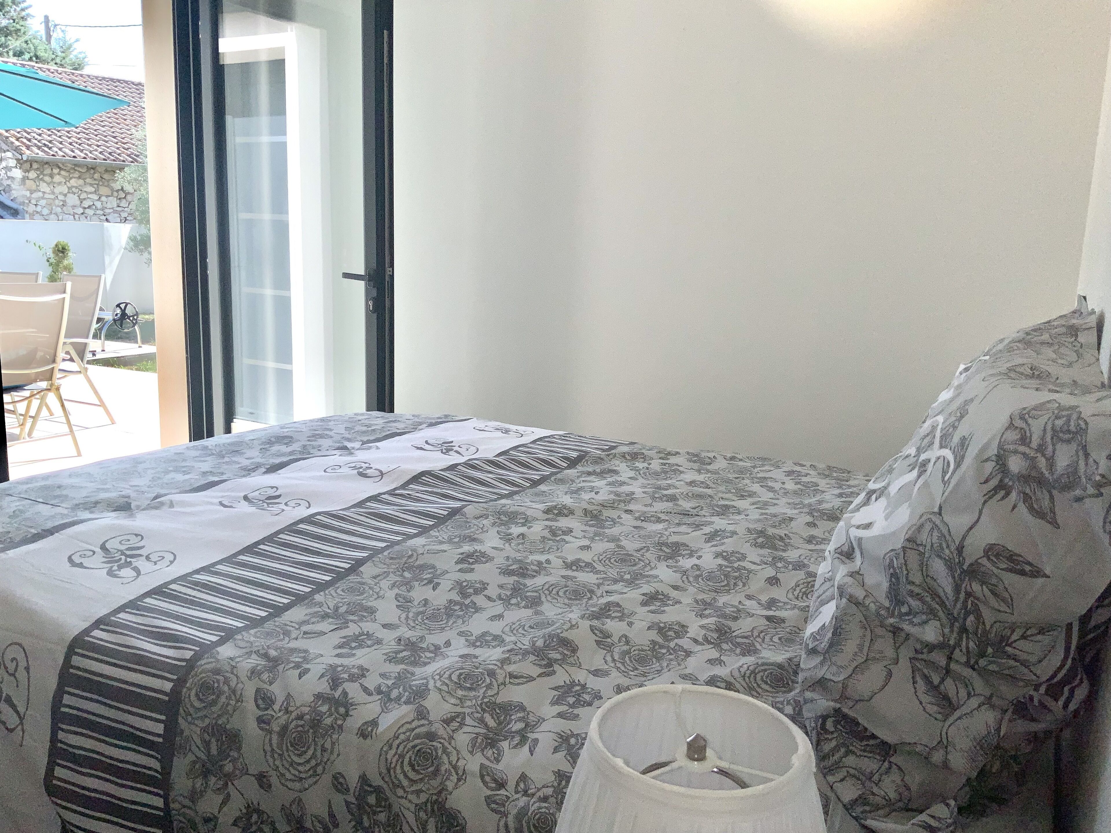 A bedroom with a bed covered in floral-patterned bedding, a bedside lamp, and a view of the outside through an open door.