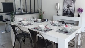 A modern dining area with a long table set for four, surrounded by dark chairs. A large mirror and a framed artwork are visible on the walls.