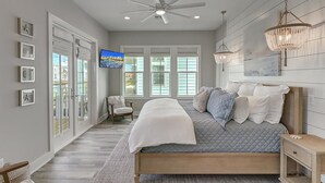 A bedroom with a large bed, a ceiling fan, a TV, and a view of the outdoors through large windows.