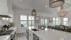 A modern kitchen with white cabinetry, stainless steel appliances, and a large island. There are pendant lights, a view through the windows, and a dining area with a bench.
