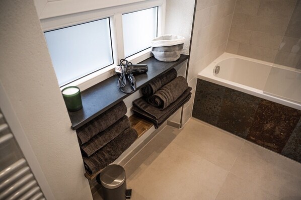A bathroom with a bathtub, a window, a shelf with towels, and a trash bin. - Rheinfreunde Bingen das Ferienhaus für Viele (Bingen am Rhein)