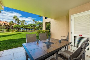 A patio area with a table and chairs, a view of a well-maintained lawn and palm trees, and a building with a beige exterior.