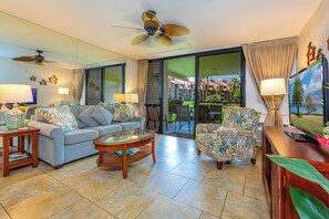 A spacious living room with a sofa, armchairs, a coffee table, and a TV. There is a balcony with a view of buildings and greenery. - Kamaole Sands #1-105 Renovated Ground Floor Unit, Partial Oceanview, A/C! (Kihei)