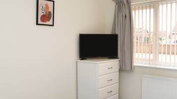 A bedroom with a bed, a TV on a dresser, a window with blinds, and a radiator.