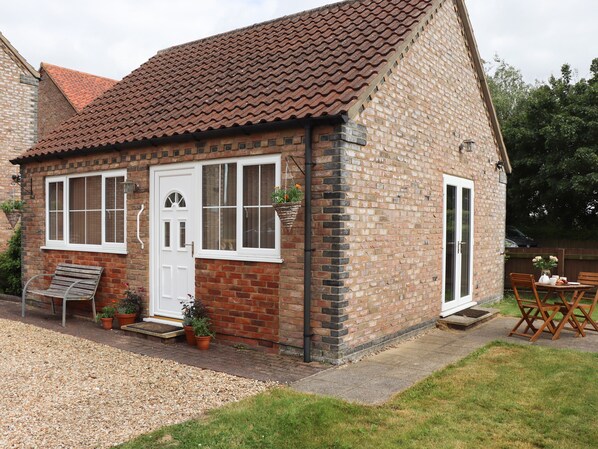 A brick cottage with a white door, two windows, a bench, and a table with chairs outside. - Copse View (Lincoln)