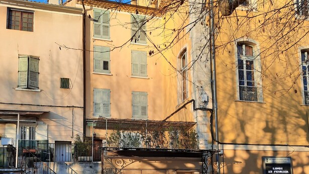 A street view with a building featuring a large wooden door, a staircase, and a signboard with text. - Typical Provencal village house with lovely terrace. (Riez)