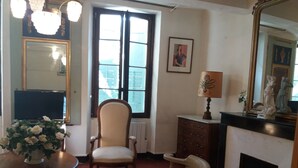A room with a large window, a chair, a table with a vase of flowers, a dresser, a lamp, and a fireplace. - Typical Provencal village house with lovely terrace. (Riez)