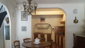 A dining area with a round wooden table, chairs, a chandelier, and a kitchen in the background. - Typical Provencal village house with lovely terrace. (Riez)