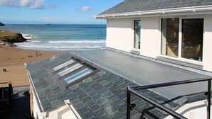 A coastal house with a sloped roof, large windows, and a view of the beach and ocean. - The White House (Wadebridge)