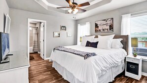 Une chambre à coucher comprenant un lit, un ventilateur de plafond, une table de chevet, un téléviseur et une porte donnant sur une salle de bain.