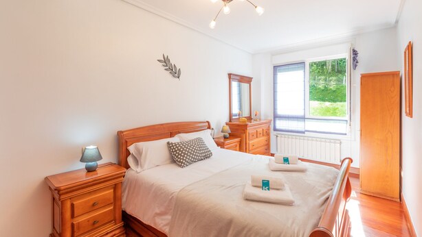 A bedroom with a wooden bed, bedside tables, a dresser, a mirror, and a window with a view of greenery.