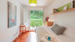 A cozy room with a bed, a small table, and a chair. There's a balcony with a view of greenery.