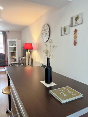 Een moderne woonkamer met een houten eettafel, een zwarte vaas met bloemen, een rode lamp en een wandklok.