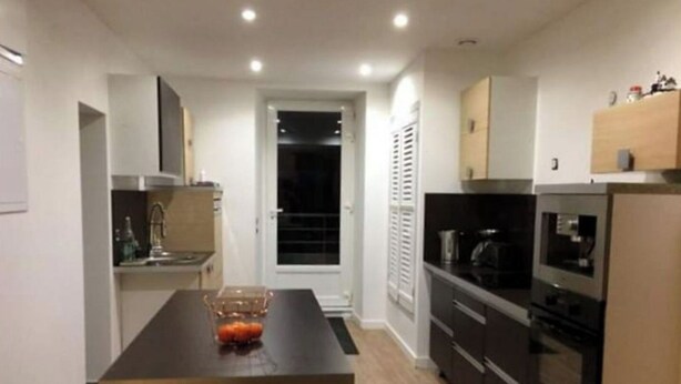 A modern kitchen with a central island, built-in oven, and a dining area with a bowl of fruit. - La Dolce Vitae (Rodez)