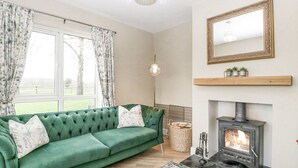 A green sofa with floral cushions, a fireplace, a mirror, and a view of a green field through a window with floral curtains. - Orchard House (Leominster)