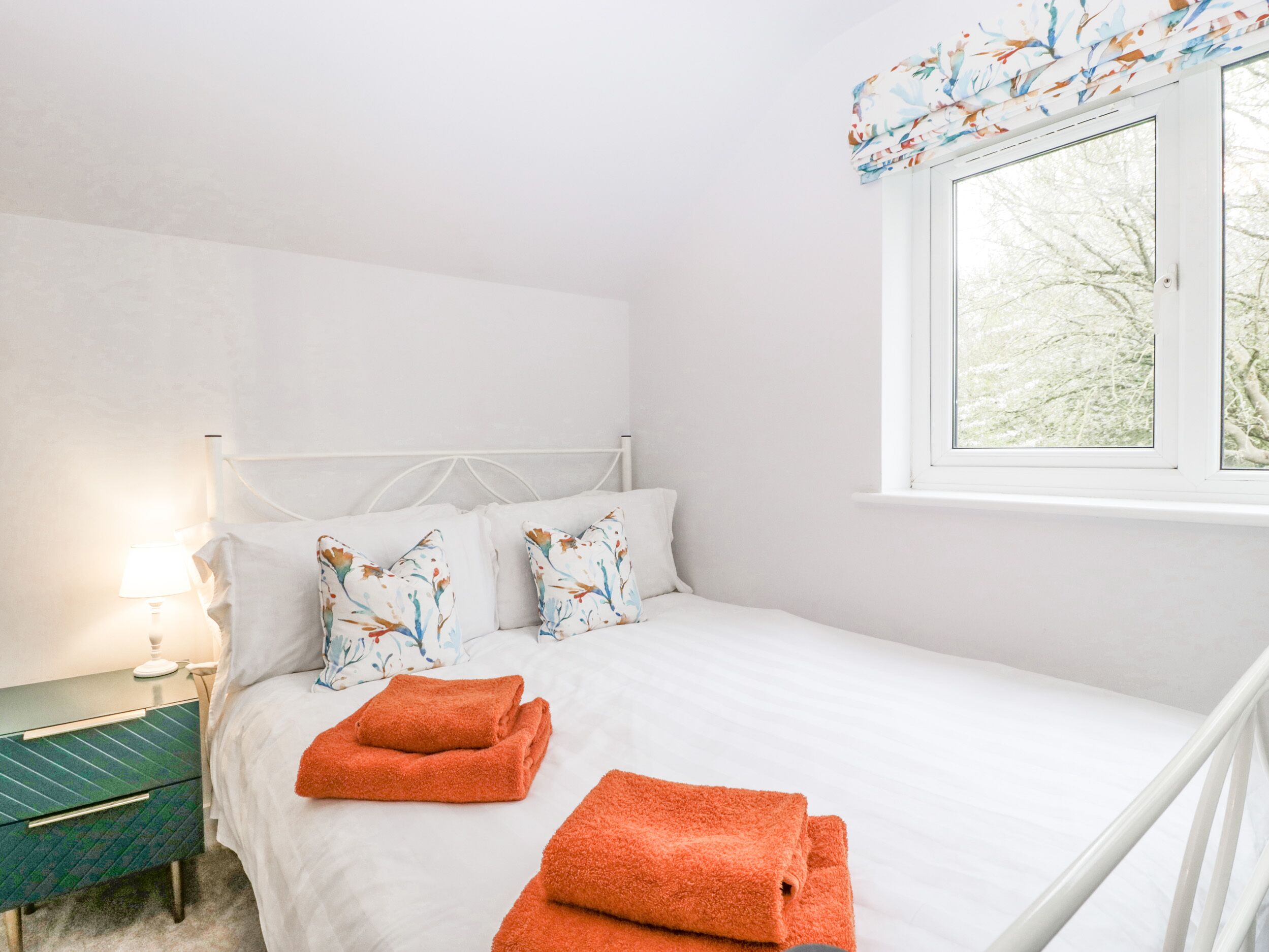 A bedroom with a bed, pillows, a nightstand, a lamp, and a window with a view of trees.