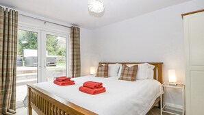 A hotel room with a wooden bed, white bedding, plaid pillows, a bedside table with a lamp, and a view of the outdoors through a sliding door. - Orchard House (Leominster)