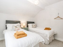Habitación de hotel con dos camas, cada una con ropa de cama blanca y almohadas decorativas. Hay una mesita de noche con una lámpara y una pequeña mesita auxiliar verde. Un perchero de madera está montado en la pared.