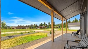 Een overdekt buitengebied met houten vloeren en Adirondack-stoelen, met uitzicht op een grasrijk landschap en woongebouwen.