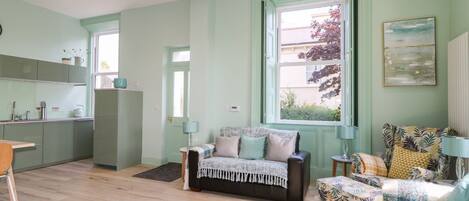 A cozy living room with a sofa and a chair, a kitchen with green cabinets, and a large window with a view of the outside.