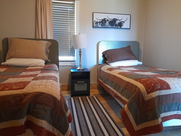 Een slaapkamer met twee bedden, een nachtkastje met een lamp, een raam met zonwering, en een aan de muur hangend schilderij.