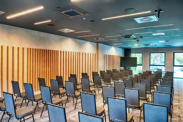 Une salle de conférence avec des rangées de chaises, un mur recouvert de panneaux en bois et des luminaires au plafond.