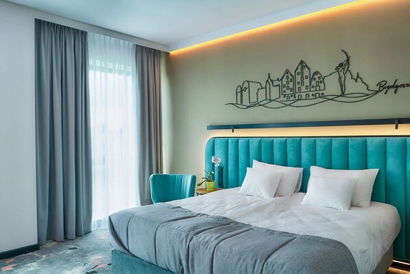 Ein Hotelzimmer mit einem großen Bett, einem dunkelgrünen gepolsterten Kopfteil, Stadtbild-Wandkunst und Blick auf die Stadt durch das Fenster.