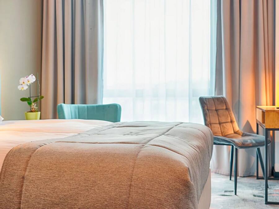 A hotel room with a large bed, a desk with a lamp, a chair, and a potted plant.