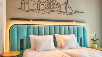 A hotel room with a teal upholstered headboard, white bedding, a cityscape wall art, a small table with a plant, and a bedside table with a lamp.