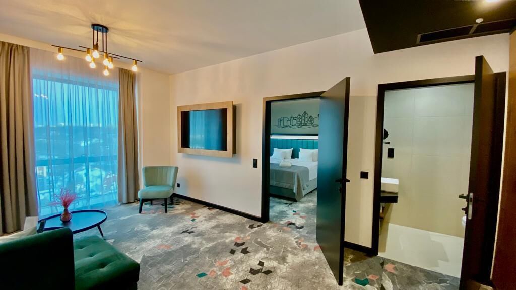A hotel room with a bed, a chair, a small table, and a view of the city.