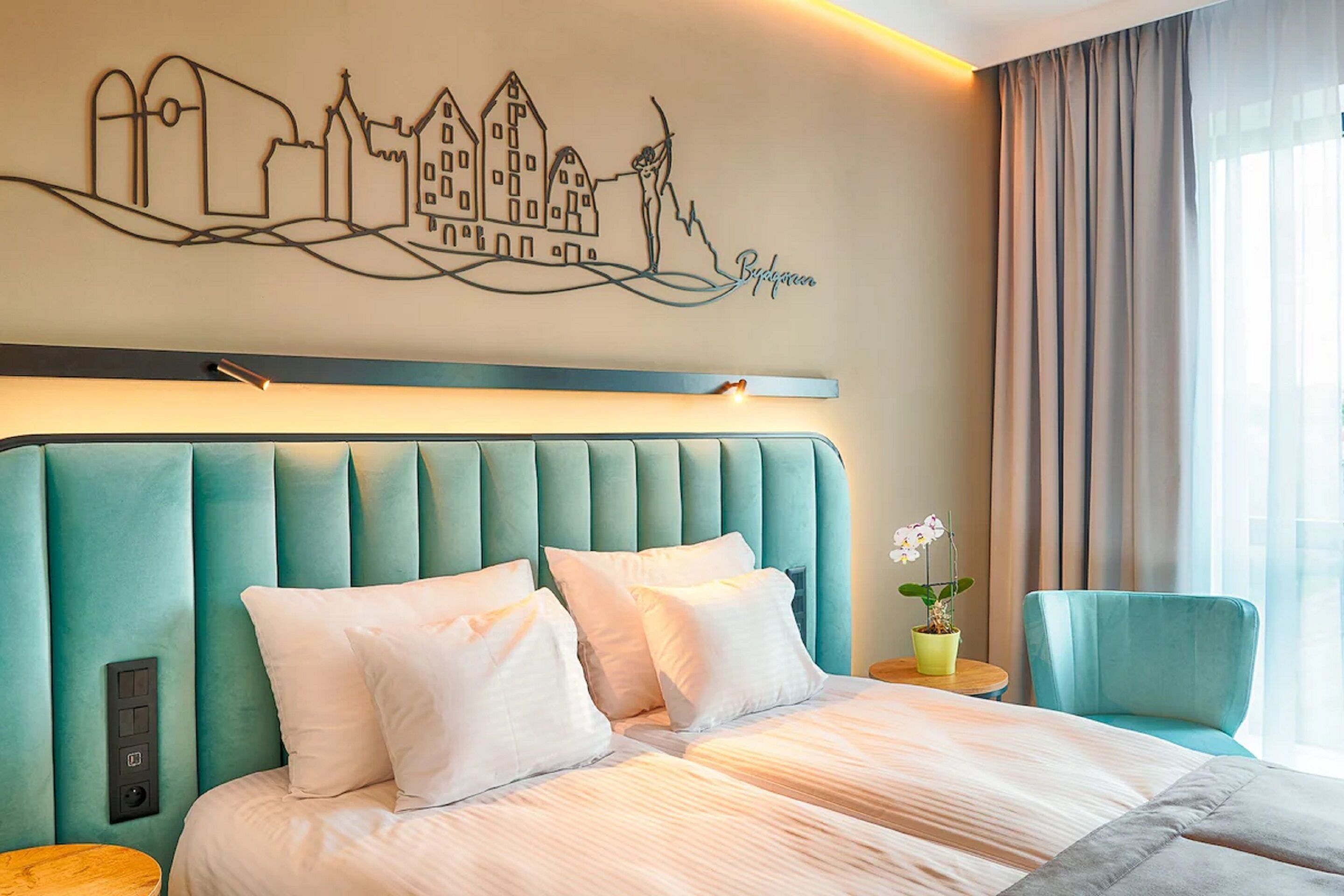 Ein Hotelzimmer mit einem großen Bett, einem Kopfteil mit Stadtbild-Fresko, einer Zimmerpflanze und einem Fenster mit Vorhängen.