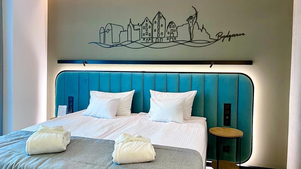 A hotel room with a teal upholstered headboard, a cityscape mural, and two neatly folded towels on the bed.