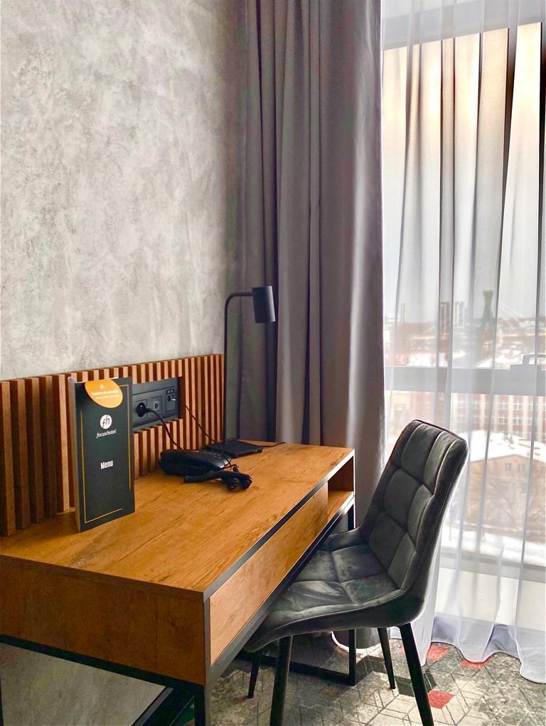 Ett modernt hotellrum med ett skrivbord i trä, en svart stol, en lampa och utsikt över staden genom tunna, genomskinliga gardiner.