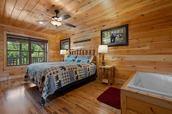 A wooden cabin bedroom with a bed, a ceiling fan, a large window, a framed picture, and a bathtub.