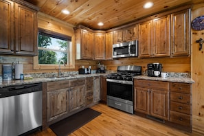 A kitchen with wooden cabinets, stainless steel appliances, granite countertops, and a window with a view of trees.
