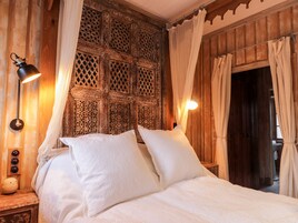 Una cama con sábanas blancas, cabecera de madera con intrincados grabados y mesita de noche con una lámpara y un objeto decorativo pequeño.