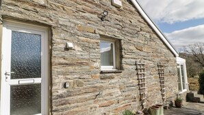 A stone building with a door, a window, and a small window above it. There are potted plants and a ladder on the ground. - The Old Smithy Cottage (Blaenau Ffestiniog)