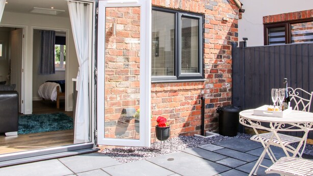 A brick house with a sliding glass door leading to a bedroom. There is a patio with a table and chairs, and a small garden area with a flower pot. - Little Oakleigh (Llanymynech)