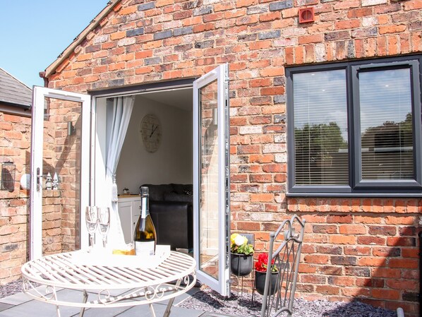 A brick house with an open door leading to a room with a white table, a bottle, and two hanging flower pots. - Little Oakleigh (Llanymynech)