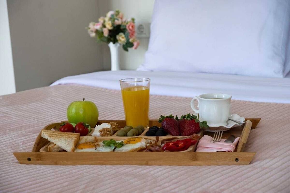 Ein Frühstücksbrett mit Toast, Eiern, Erdbeeren, einem grünen Apfel und einem Glas Orangensaft auf einem Bett mit einem weißen Kissen und einem Blumenstrauß im Hintergrund.