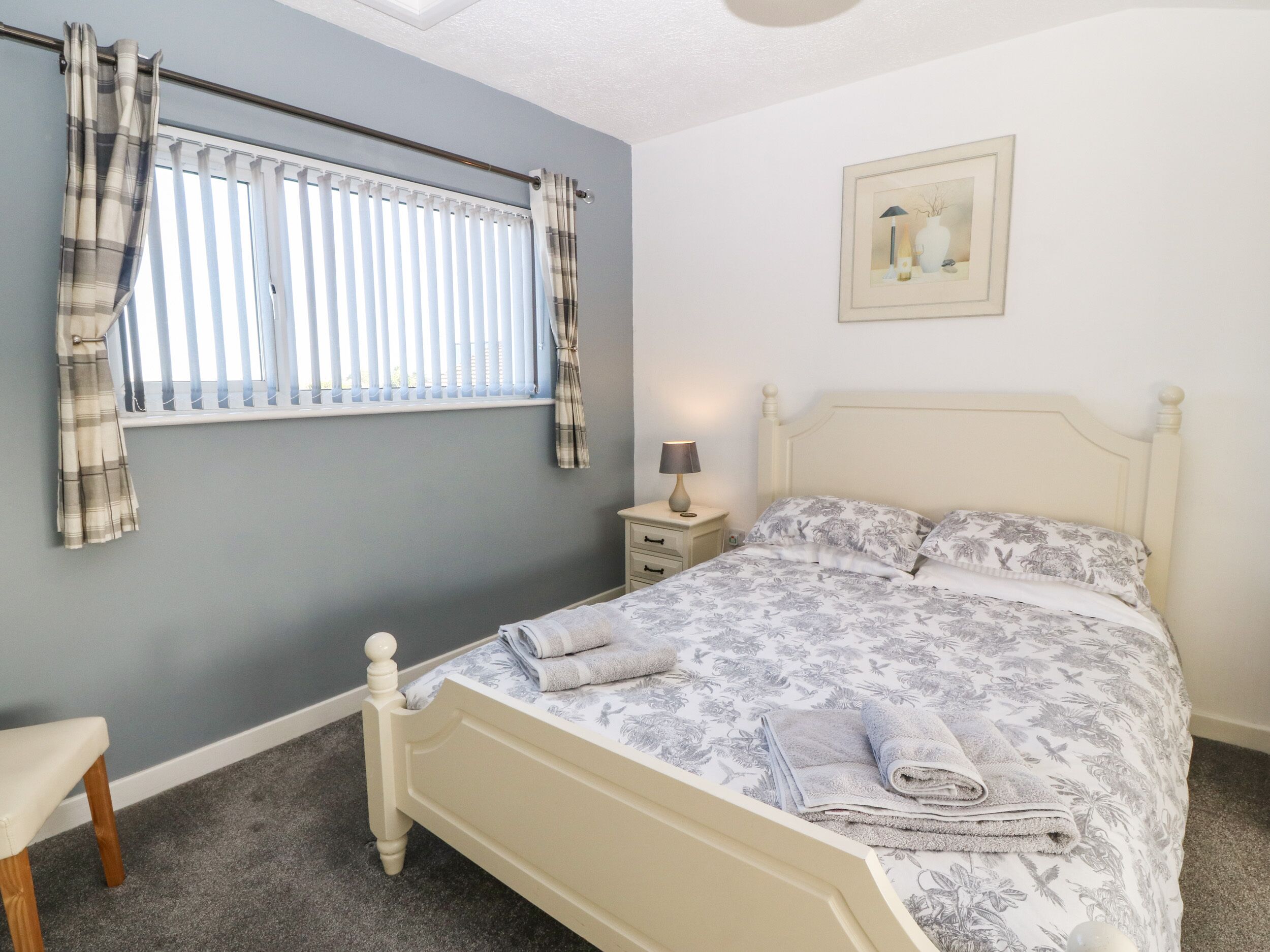 Un dormitorio con cama, mesita de noche, lámpara, ventana con cortinas e imagen enmarcada en la pared.